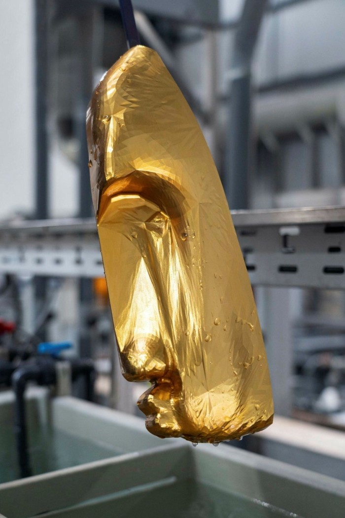 A long golden sculpture resembling an ancient mask hangs in an industrial gallery space 