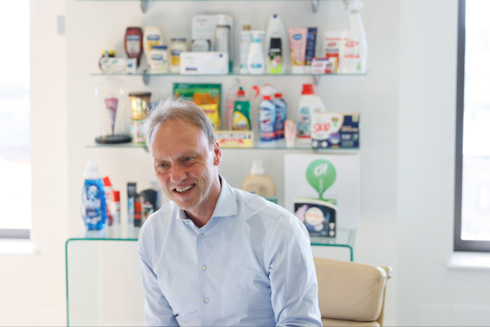 A smiling middle-aged Caucasian man in a light blue shirt in front of shelves stocked with various household cleaning products