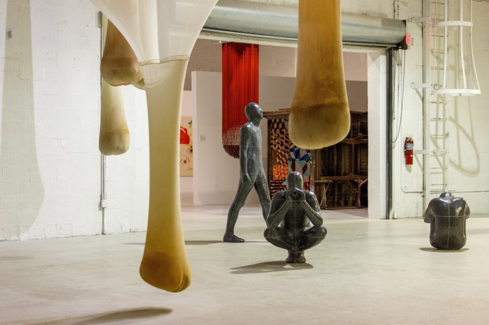 View of a gallery in a warehouse with several artworks: three statues of human figures, brown translucent sacks stretching down from the ceiling, abstract painting and more installations in the background