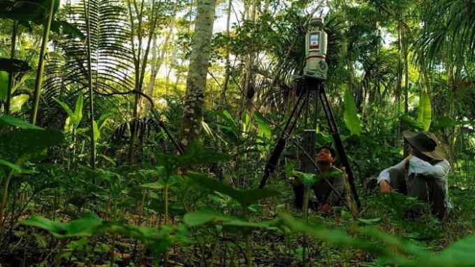 Researchers sit by a tripod-mounted monitor in a rainforest