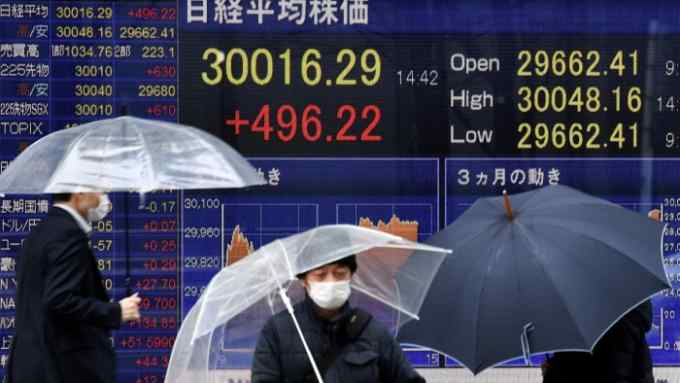 Pedestrians holding umbrellas walking past an electronic quotation board that displays share prices