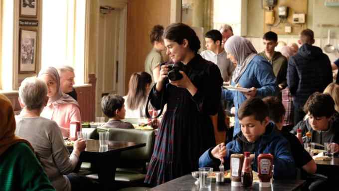 A smiling woman carrying a camera stands in the middle of a room where adults and children are sitting at tables, eating and drinking