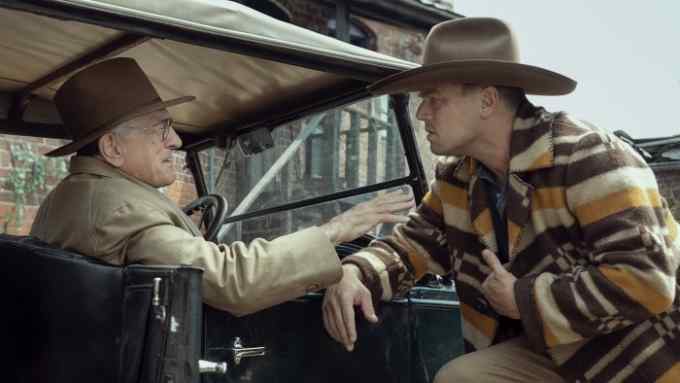 An older man wearing a hat sits at the wheel of a vehicle of the 1920s talking to a younger man who wears a hat and a jacket decorated in horizontal stripes