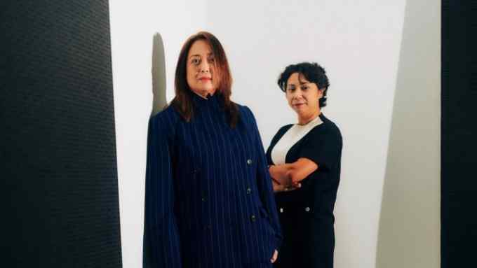 Two women, one in a blue pinstripe suit, the other in a black and white top, look seriously at the camera