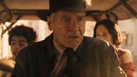 A man in a fedora rides in a vehicle looking anxious; behind him are a boy and a woman