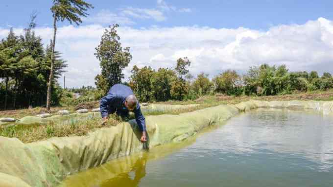 A man inspects a large water pan, lined with plastic sheet, in the middle of a grassland