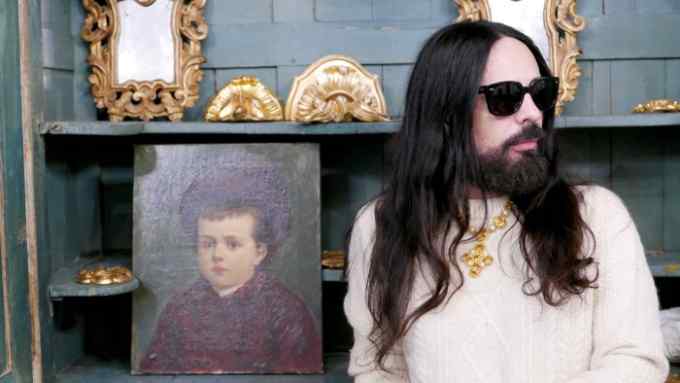 Alessandro Michele, creative director of Gucci, with dark eyeglasses and posing beside a portrait of a child