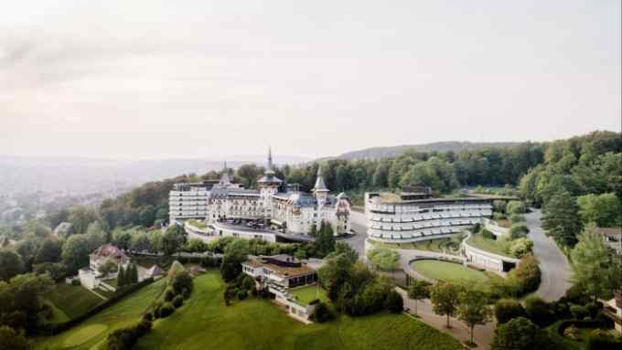 An aerial shot of Zürich’s Dolder Grand hotel: a white, turreted late-19th-century building flanked by two curving, modernist wings, sitting on a forested hill overlooking the city