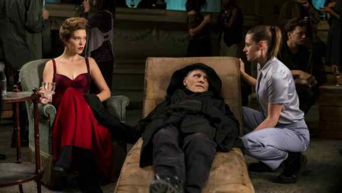 A woman in a red silk dress sits in an armchair next to a man in black on a chaise longue, who is talking to a woman in grey