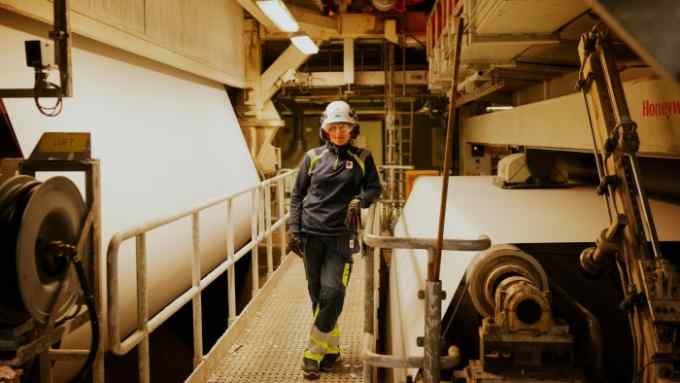 A woman in her work clothes and safety gear poses for the camera inside a pulp and paper mill
