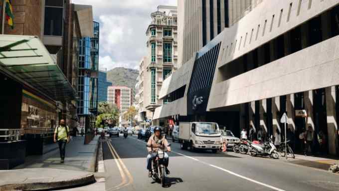 A snapshot of a street in Port Louis, Mauritius. There is a motorcycle rider, a pedestrian on the sidewalk, plus cars and buildings in the background