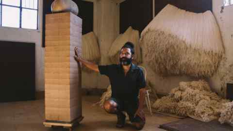 A bearded man wearing a black shirt and shorts kneels next to a sculpture with piles of fabric threads behind him