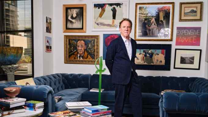 A middle-aged man wearing a blue suit and a white shirt stands in his art-and-books-filled office in front of a velvet blue couch