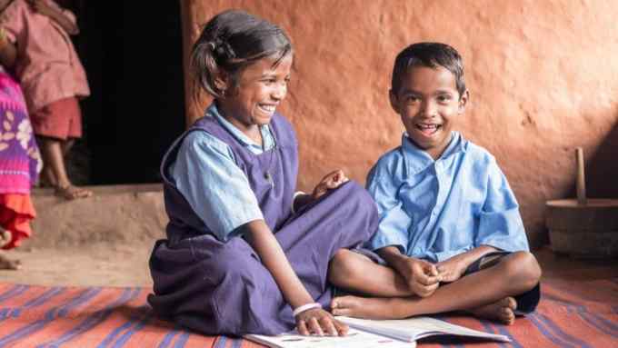 Gumeshwari and Kiran study at home after school in Tirathgarh Village, Bastar, Chattisgarh, India. Two children in school uniforms are sitting and laughing on a colorful mat outside a rural house with mud walls, while a woman holding a child looks on with a smile from the shadowed doorway