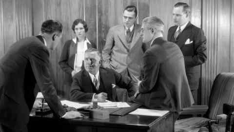 1930s WORRIED BUSINESS GROUP FIVE MEN AND ONE WOMAN IN OFFICE MEETING AROUND THE BOSS’ DESK