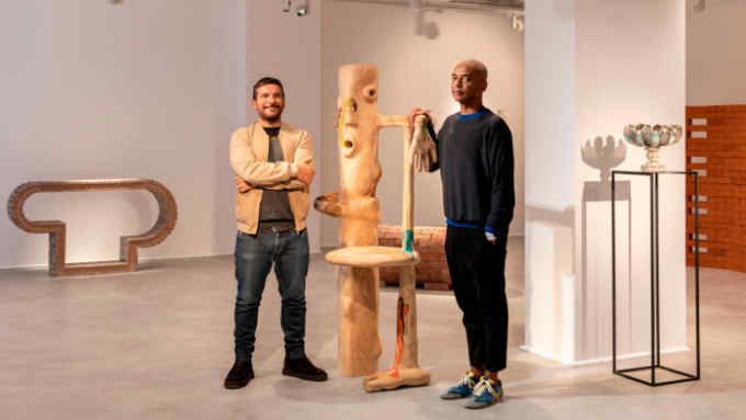 Two men standing in a large white gallery either side of a wooden sculpture which looks like a bare tree trunk with a face