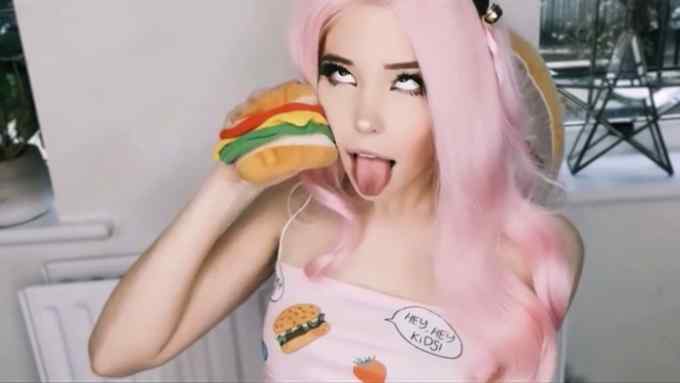 A young woman in a pink wig pulls a face for the camera, holding a toy hamburger to her ear