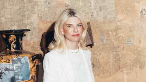 A glamorous blond woman in a white jacket and brown trousers
