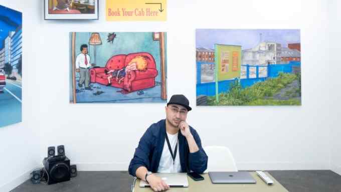 A man with glasses in a black baseball cap and navy baseball jacket sits at a table in front of colourful paintings