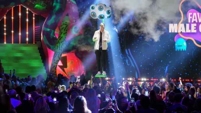 The image shows an energetic and vibrant scene from an award show or entertainment event. A man stands on stage, highlighted by a spotlight, in front of a colorful backdrop with green neon lights