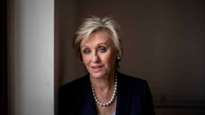 Tina Brown in smart attire leans against a wall next to a window