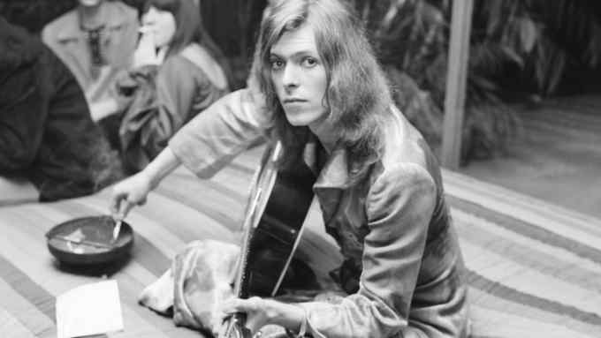 A young man with long hair sits holding a guitar while flicking ash from a cigarette into an ashtray