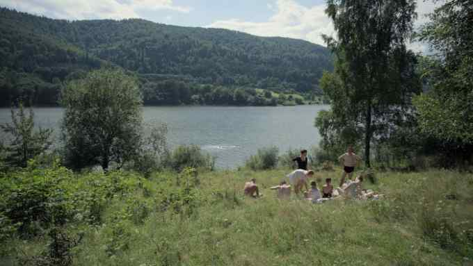People in 1940s swimming costumes sit in long grass nexst to a lake