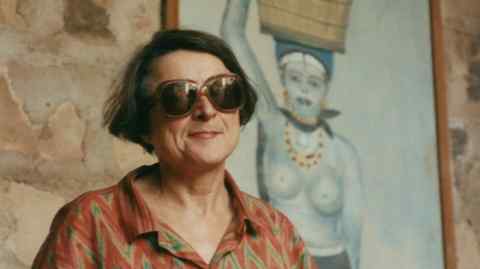 A woman in sunglasses looks cool in front of a blue painting of a bare-breasted woman