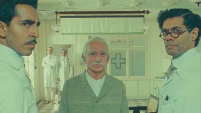 In a medical-looking environment, three men stand together looking slightly puzzled; in the background are two figures in white medical clothing