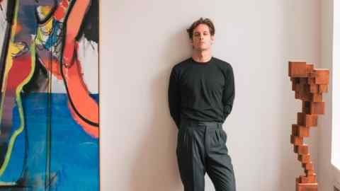 A tall man with thick wavy hair and wearing a dark grey sweater and trousers stands against a wall next to a sculpture and an abstract painting