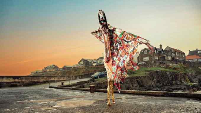 A person on stilts wearing bright flowing clothes and a head dress standing in a seaside village at dusk