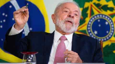 A man in his seventies and in a suit, with Brazil flags on the wall behind him, gesticulates as he talks
