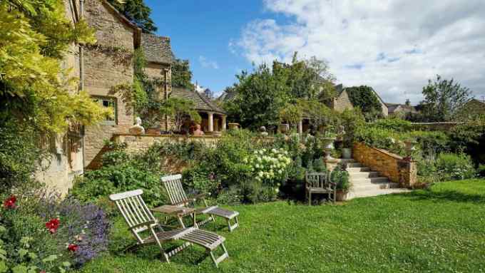 A stone house with a lush garden outside. A table and two outdoor seats for lying down are in the garden