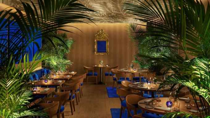 The palm-filled, blond-wood interior of The Rome Edition’s restaurant, with, its floor dotted with indigo rugs