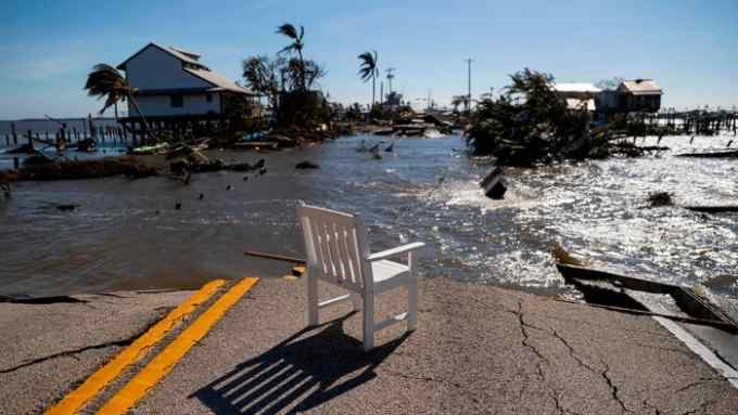 A garden chair standing in the middle of a road - the rest of the road in the back has been washed away by floods, with houses and palm trees in the background