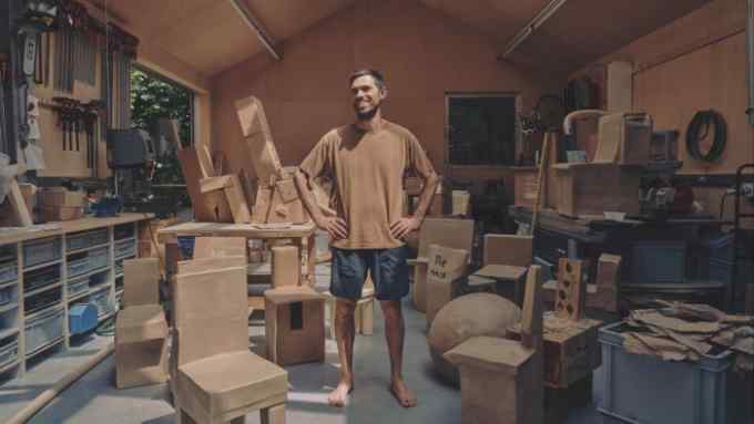 A man in a brown T-shirt stands in his wooden studio surrounded by cardboard furniture in the same shade of brown