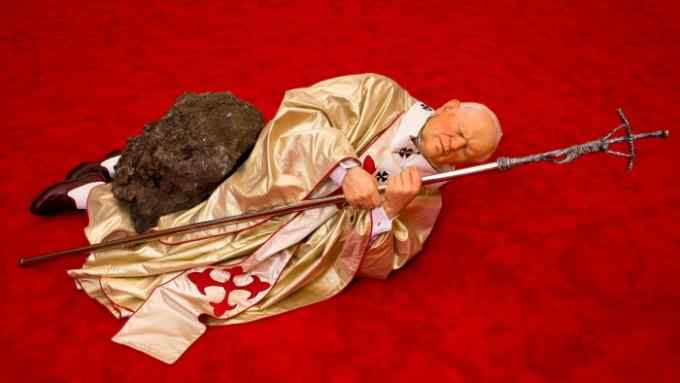 A lifelike sculpture of Pope John Paul II in golden robes, clutching a sacred staff, lying on the ground, hit by a meteorite