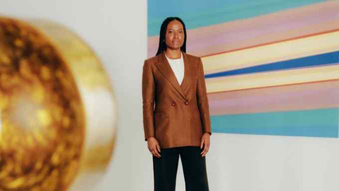 Photograph of a woman in a brown blazer and dark trousers, standing between a painting and a sculpture, taken in a gallery