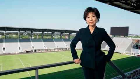 An Asian woman in a dark suit stands in the terrace of a football stadium
