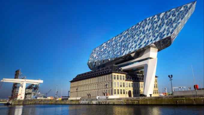 The building has a diamond-shaped patterned facade and an angular, cantilevered shape that overhangs the base. The building is situated by a body of water with a clear blue sky above