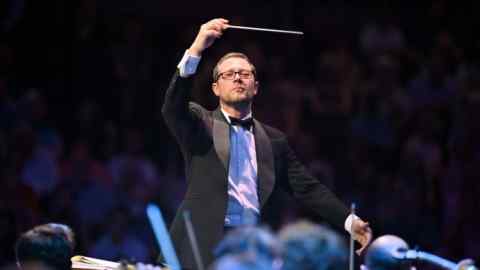 A male conductor in a black-tie suit raises his baton elegantly in front of an orchestra