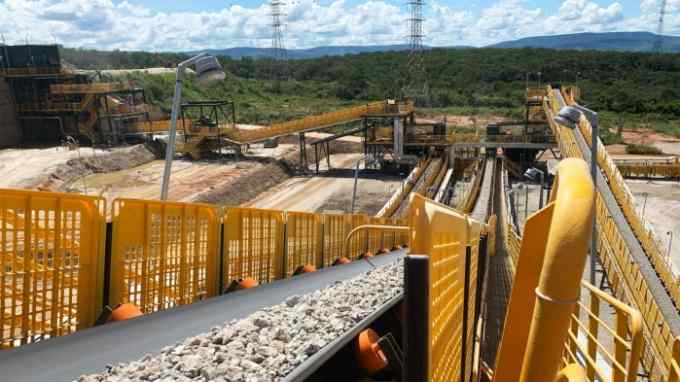 A conveyor belt loaded with gravel running through a large industrial mining site with various machinery and structures in the background, all secured by bright yellow safety barriers