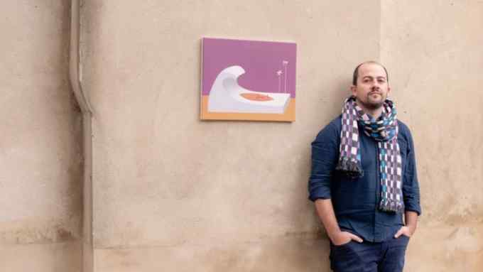 A man in blue shirt and jeans leans against a putty-coloured wall with a painting of a white wave against a pink sky