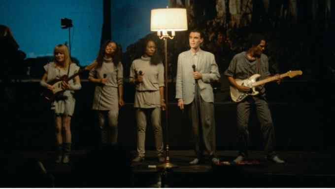 A group of musicians stand on a stage illuminated by a large floor lamp; two are playing guitars