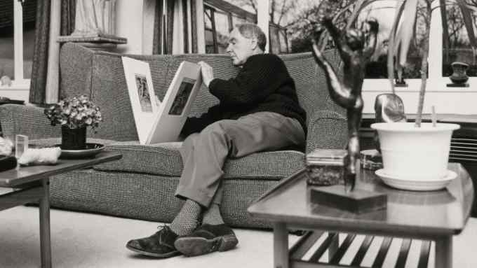 An elderly gentleman engrossed in a large art book while sitting on a textured sofa, with artistic objects and furniture in the foreground