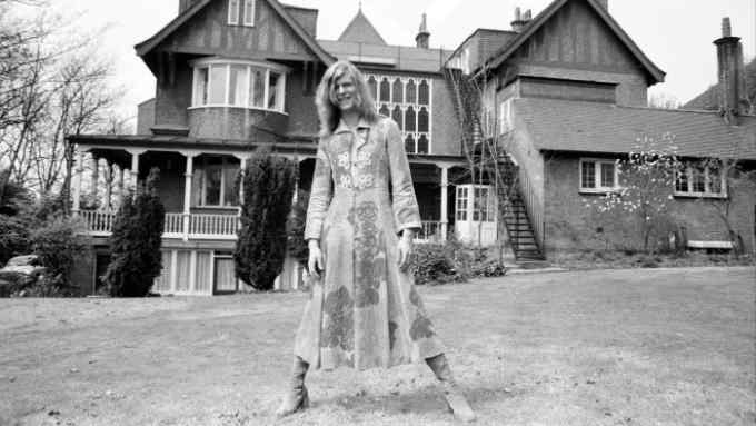 A man wearing a dress stands on the lawn outside a large rambling house