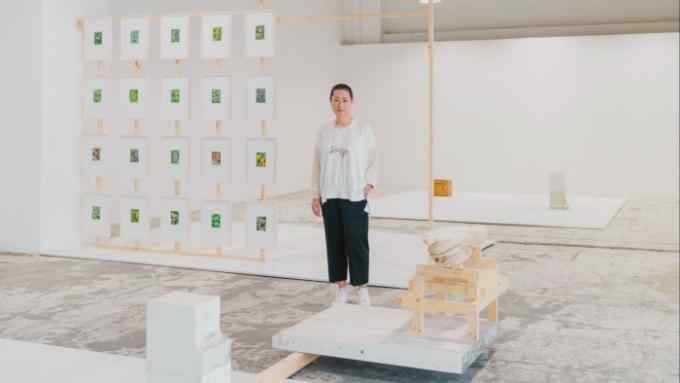 A woman with short hair and glasses worn on a chain around her neck stands in a minimalist gallery space with a concrete floor