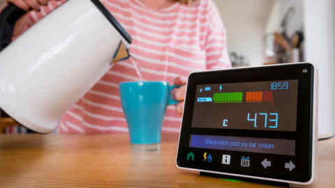 A household electricity monitor with a woman making a cup of tea in the background