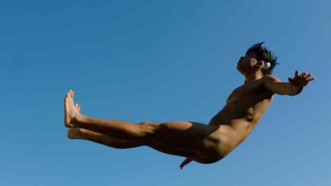 A young naked man is captured  falling backwards mid-air against a clear blue sky.
