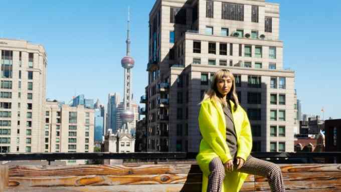 A person in a long neon-yellow coat sits on the wooden edge of a rooftop in front of skyscrapers